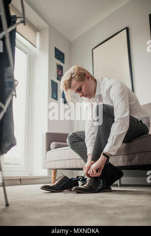 Low angle view of a young businessman sitting on the sofa tying his shoe laces. Stock Photo
