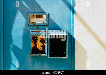 Rusty mailboxes and a mail slot on a blue door next to a white wall. Stock Photo