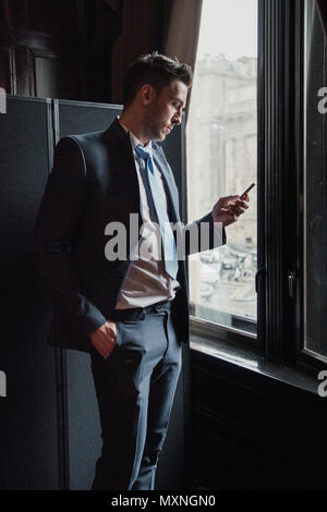 Side view of a young businessman standing next to a window in the office. He is checking his smart phone while waiting. Stock Photo