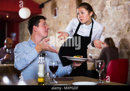 Dissatisfied male expressing displeasure with food talking to excusing waitress in restaurant. Focus on man Stock Photo