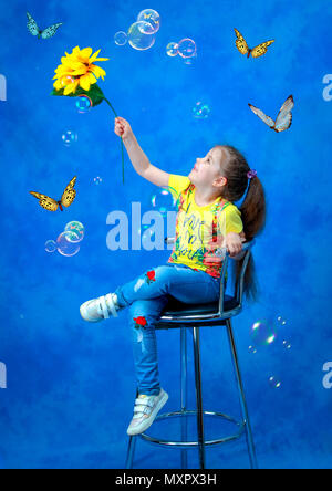 Studio shooting. A girl with a sunflower in her hand on a blue background catches butterflies and soap bubbles. Stock Photo