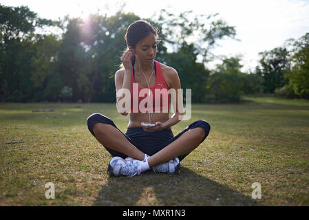 Young atheletic woman sitting on the grass in a field getting after a run in the park holding her mobile phone with ear buds in listening to music Stock Photo