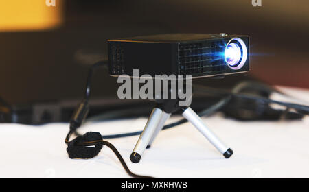 Black projector with tripod installed on white table, in hall or seminar meeting room. Real projector at business conference or presentation. Stock Photo