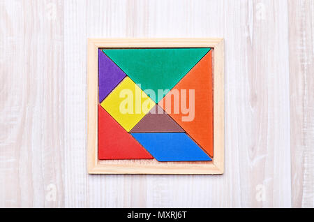 A tangram puzzle consisting of colored pieces with geometric shapes, collected in a square on a light wooden background Stock Photo