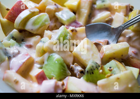 Plate with chopped apples and kiwis mixed with granola and yogurt. A steel spoon rest in the fruit. Stock Photo