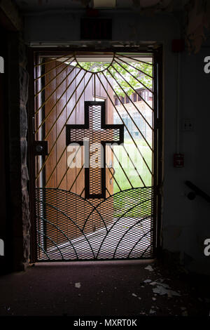 Light coming through metal gate with cross from inside church in prison yard with razor wire. Stock Photo