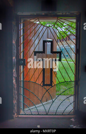 Light coming through metal gate with cross from inside church in prison yard with razor wire. Stock Photo