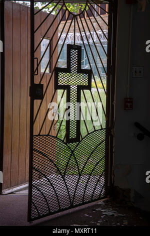 Light coming through metal gate with cross from inside church in prison yard with razor wire. Stock Photo