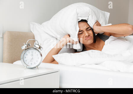 Portrait of caucasian upset woman shutting ears with pillow due to ringing alarm clock, while lying in bed Stock Photo