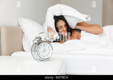 Portrait of annoyed young woman covering ears with pillow due to focused ringing alarm clock in morning while lying in bed Stock Photo