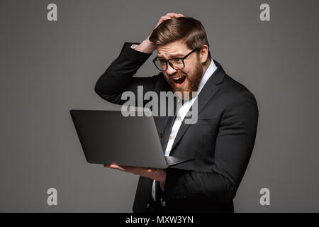 Portrait of a disappointed young businessman dressed in suit looking at laptop computer isolated over gray background Stock Photo