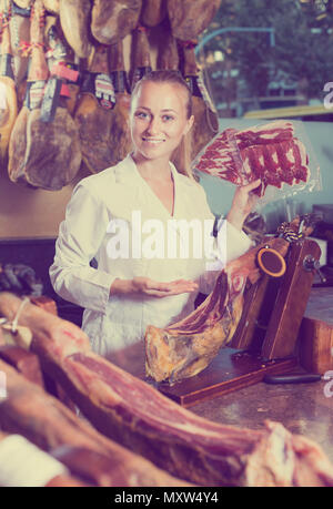 Positive young woman holding iberian ham in meat shop indoors Stock Photo