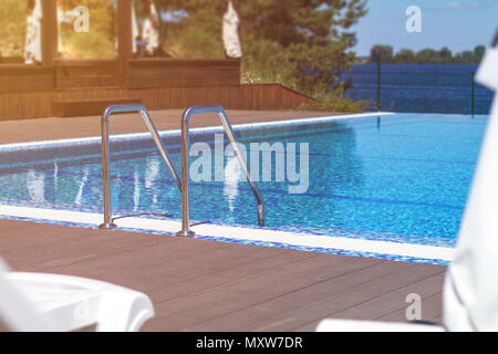 Swimming pool with stair and wooden deck at hotel. Grab bars ladder in the blue swimming pool. Summer sunny day. Stock Photo