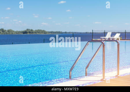 Swimming pool with stair and wooden deck at hotel. Grab bars ladder in the blue swimming pool. Summer sunny day. Stock Photo