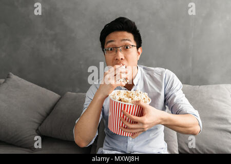 Portrait of a scared young asian man eating popcorn while sitting on a couch at home and watching TV Stock Photo