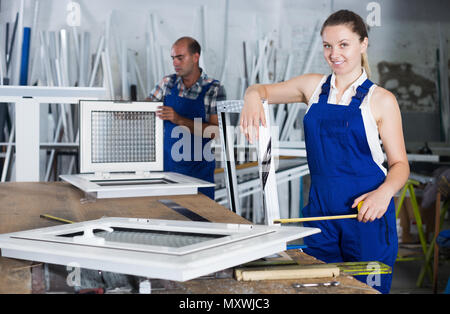 Smiling workwoman in overalls using measuring tape and standing with pvc window Stock Photo