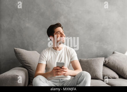 Content smiling man in basic t-shirt holding smartphone in hands and looking aside with happy gaze while sitting on sofa inside Stock Photo