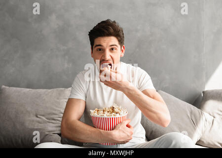 Single man 20s sitting on sofa in gray apartment and eating pop corn while watching tv in morning Stock Photo