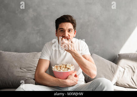 Hungry caucasian man 20s sitting on sofa in house and eating pop corn while watching on you Stock Photo