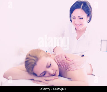 Aged massagist is doing relaxing massage shoulder area for young woman in spa salon. Stock Photo