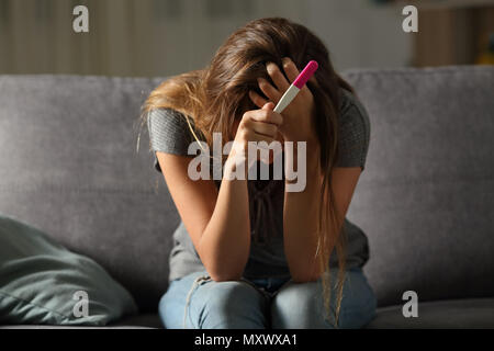 Single sad woman complaining holding a pregnancy test sitting on a couch in the living room at home Stock Photo