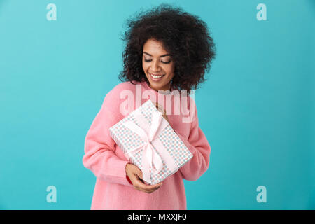 Portrait of joyous woman 20s with afro hairdo holding gift box and smiling in happiness isolated over blue background Stock Photo