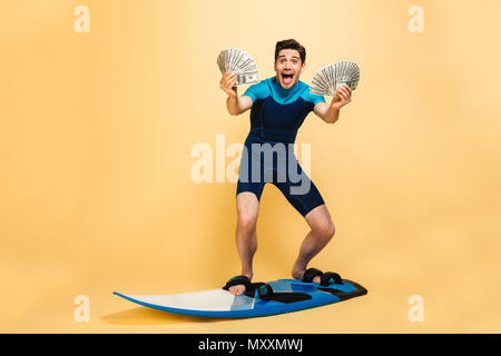 Full length portrait of an excited young man dressed in swimsuit holding bunch of money banknotes while surfing on a board isolated over yellow backgr Stock Photo