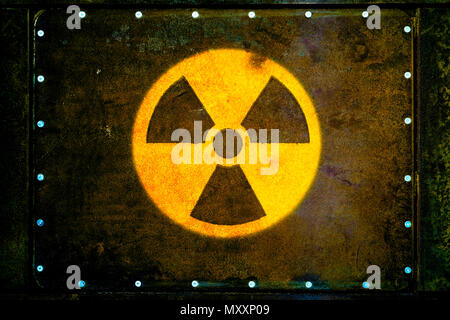 Round yellow radioactive (ionizing radiation) danger warning symbol painted on massive rusty metal plate fixed with metallic rivets to the wall with d Stock Photo