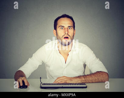 Casual man in white shirt sitting at table and using computer with expression of deep surprise Stock Photo