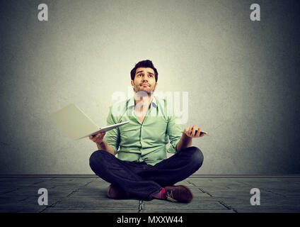 Casual man sitting on floor with laptop in hands while shrugging with shoulders in misunderstanding Stock Photo