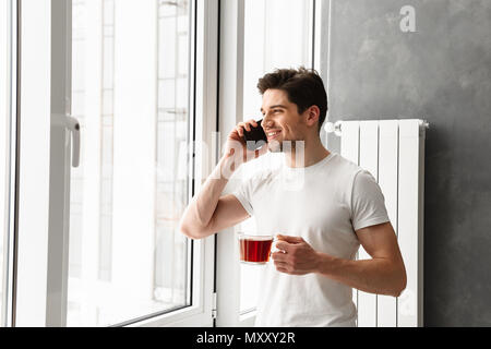 Masculine brunette man speaking on mobile phone and looking out the window while holding glass with tea Stock Photo