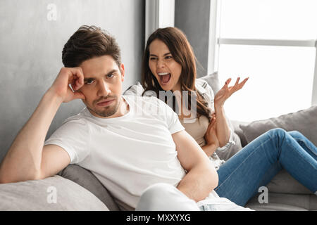 Photo of disappointed couple sitting together on sofa at home with upset look while woman screaming on man isolated over white background Stock Photo