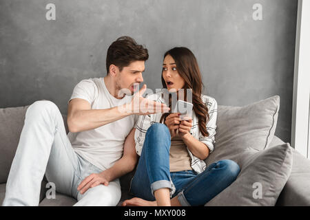 Displeased couple sitting together on sofa at home while man shouting on woman who holding smartphone in hand isolated over white background Stock Photo