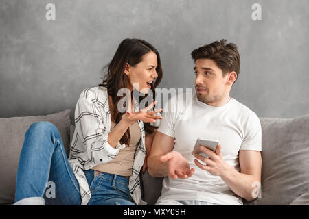 Fighting couple sitting together on sofa at home while woman shouting on man who holding smartphone in hand isolated over white background Stock Photo