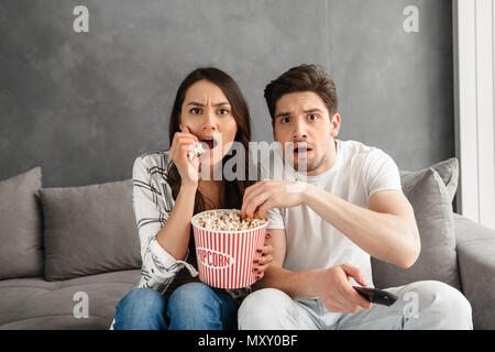 Image of anxious family sitting on sofa at home and looking at you while watching scary movie and eating popcorn Stock Photo