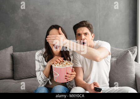 Image of caucasian uptight family sitting on sofa at home with popcorn in hand while watching scary movie Stock Photo