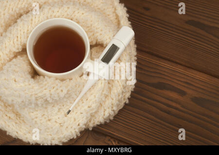 A tea mug wrapped in a soft fluffy knitted scarf, a thermometer on a wooden background - the concept of seasonal autumn diseases, colds, flu treatment Stock Photo