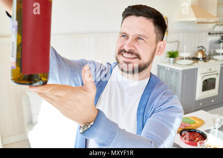 Portrait of smiling bearded man holding wine bottle, inspecting color and quality while setting dinner table  waiting for guests in sunlit dining room Stock Photo