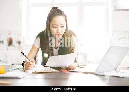 Portrait of focused creative Asian woman drawing sketches for while standing at work table in sunlit fashion design atelier, lens flare Stock Photo