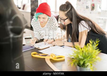 Portrait of two contemporary creative young women working in fashion design drawing sketches and collaborating on project standing by tailors table wi Stock Photo