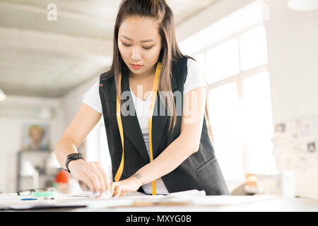 Portrait of young Asian woman  making patterns while standing at tailors table and working in modern design atelier lit by sunlight Stock Photo