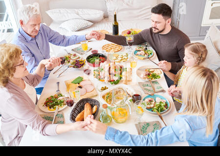 High angle portrait of big happy family joining hands in prayer at dinner holding hands during festive celebration in sunlight Stock Photo