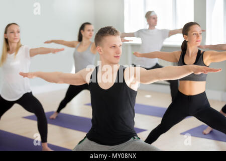 Group of young sporty people practicing yoga lesson, doing Warrior exercise, Virabhadrasana 2 pose, working out, indoor close up, yogi students traini Stock Photo