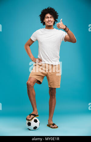 Image of happy young african curly man football player standing isolated over blue background holding ball showing thumbs up. Stock Photo