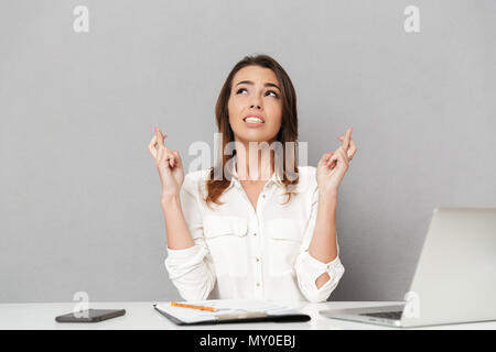 Portrait of a worried young business woman sitting at the office desk and holding fingers crossed isolated over white background Stock Photo