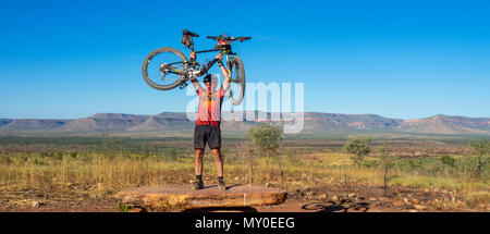 A cyclist on the Gibb Challenege 2018 holding up his bike in celebration at Cockburn Ranges lookout, Kimberley, WA, Australia. Stock Photo