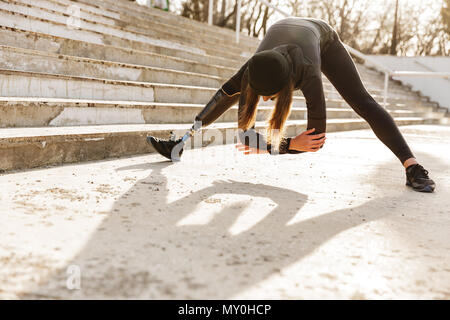 Image of disabled sportswoman in black tracksuit having prosthetic leg exercising with slopes and stretching body outdoors Stock Photo