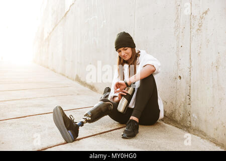 Photo of content handicapped woman having bionic leg in streetwear sitting on concrete floor outdoor and looking at wristwatch with thermos cup in han Stock Photo