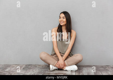 Portrait of a smiling asian woman sitting on a floor with legs crossed and looking away over gray background