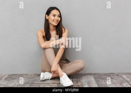 Portrait of a pretty asian woman sitting on a floor with legs crossed and looking away over gray background